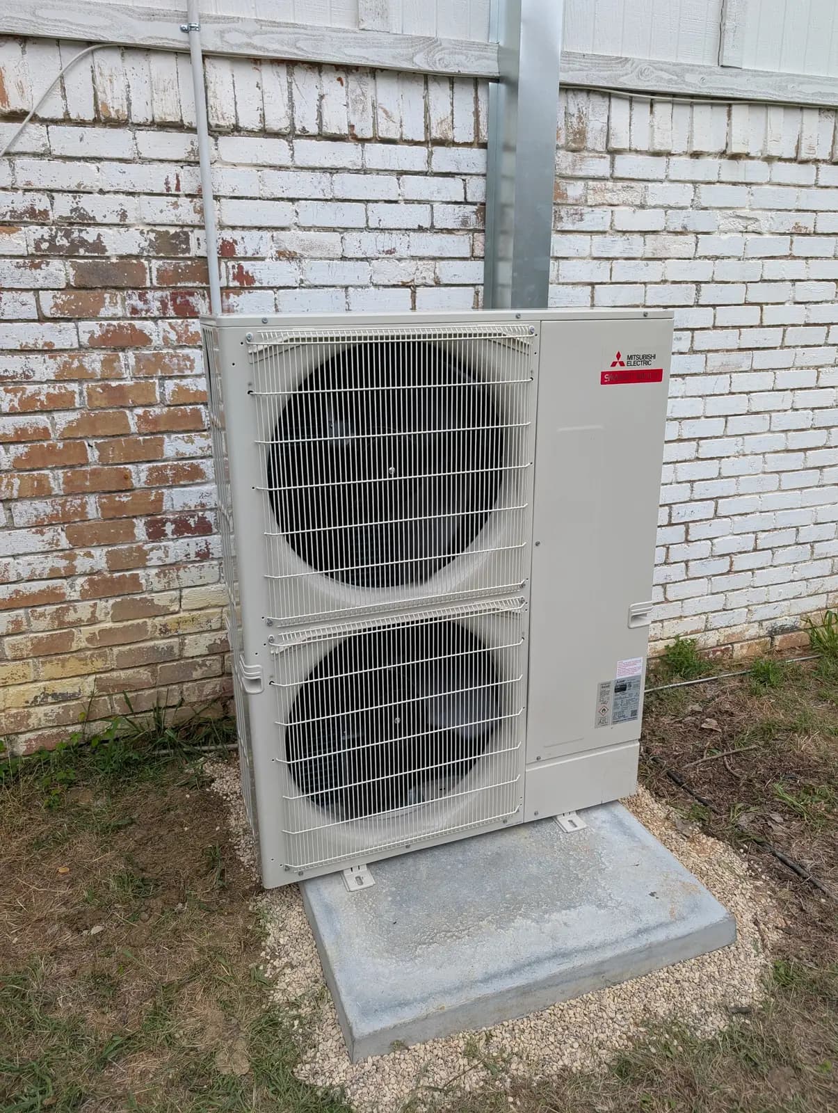 Mitsubishi Electric two-fan outdoor unit on a pad beside painted brick