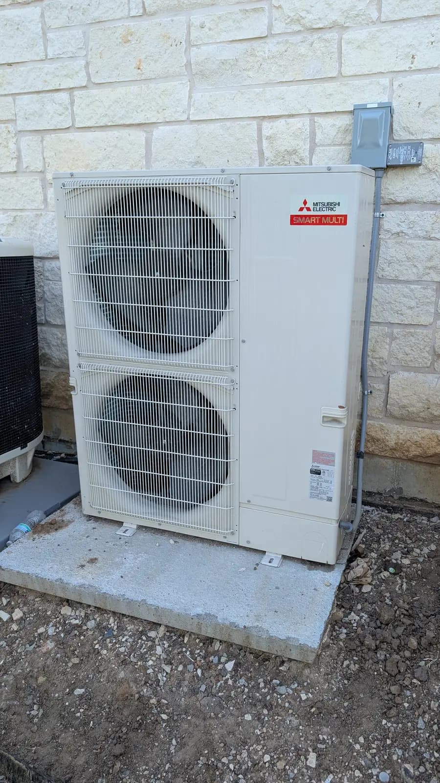 Mitsubishi Electric Hyper-Heat two-fan outdoor unit on a concrete pad by a limestone wall