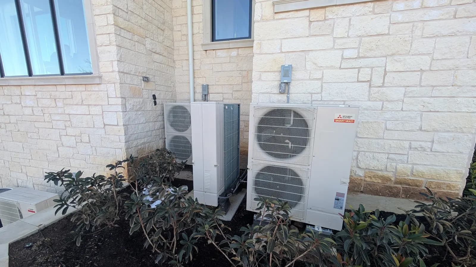 Two Mitsubishi Electric outdoor units in a landscaped bed against a limestone house