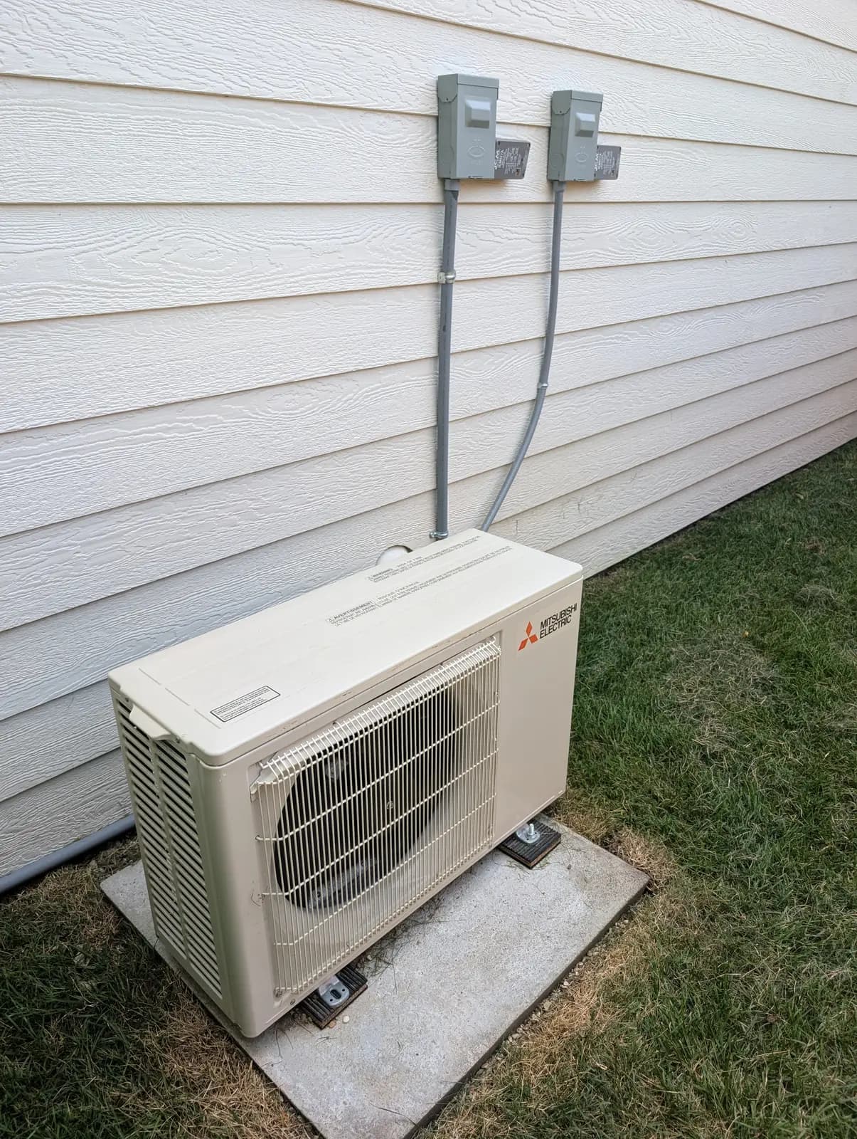 Mitsubishi outdoor unit on a pad in grass beside white lap siding