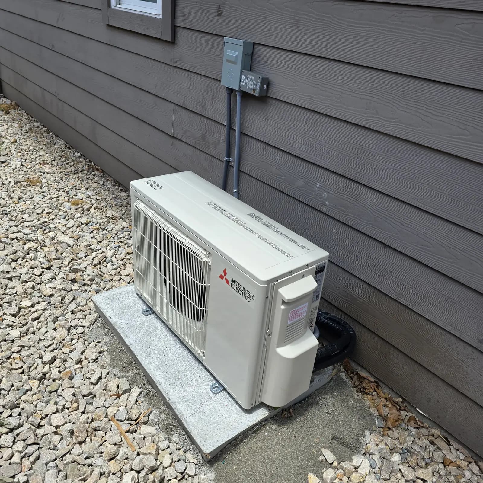 Mitsubishi Electric condenser on a pad in gravel beside dark gray lap siding
