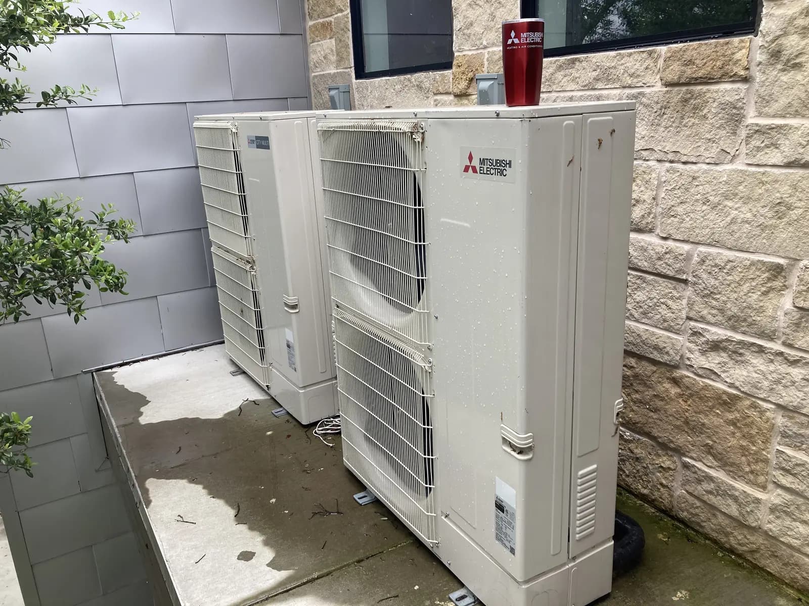 Two large Mitsubishi Electric outdoor units on a pad against a stone and metal wall