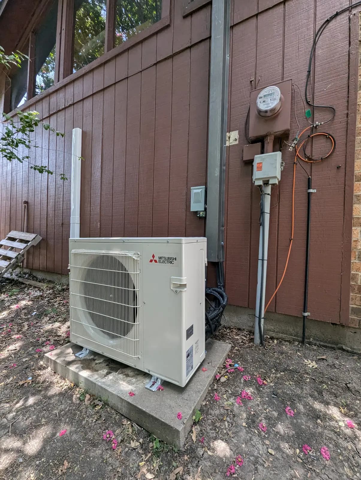 Mitsubishi Electric outdoor unit on a pad beside red-brown siding under trees