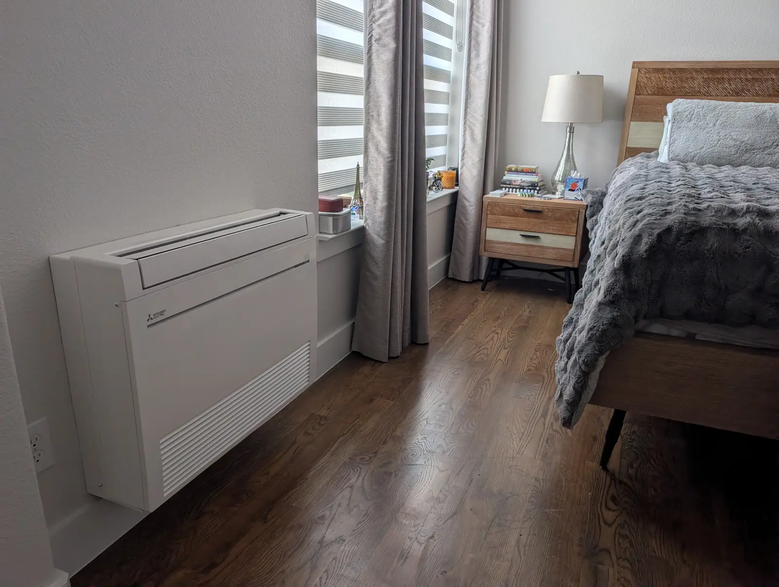Floor-mounted mini split under a window in a bedroom with wood floors and a bed