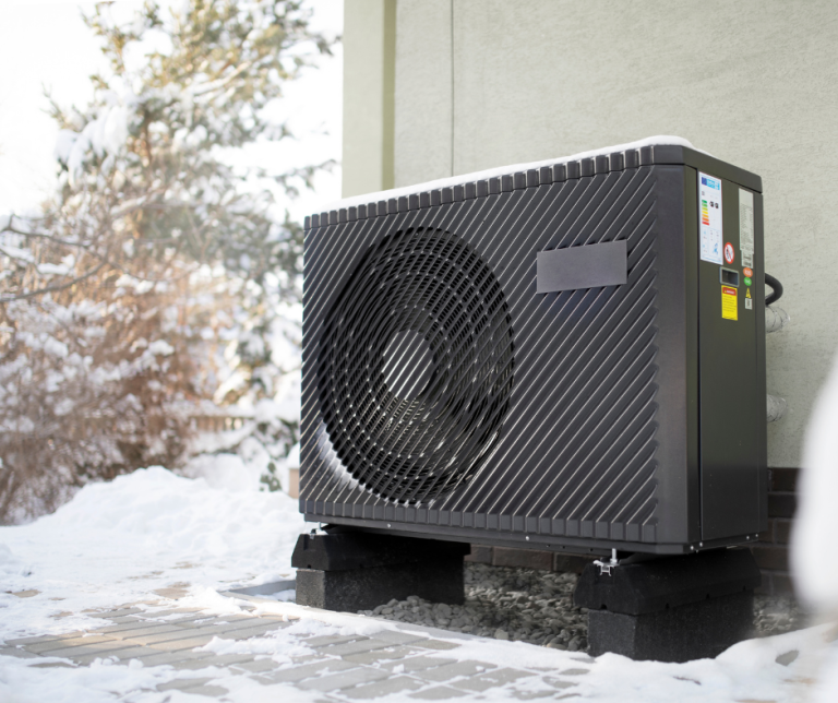 Black outdoor heat pump unit mounted on raised blocks against a white wall with snowy ground beneath. The unit features a large circular fan grille with diagonal venting patterns and energy efficiency labels visible on the side.