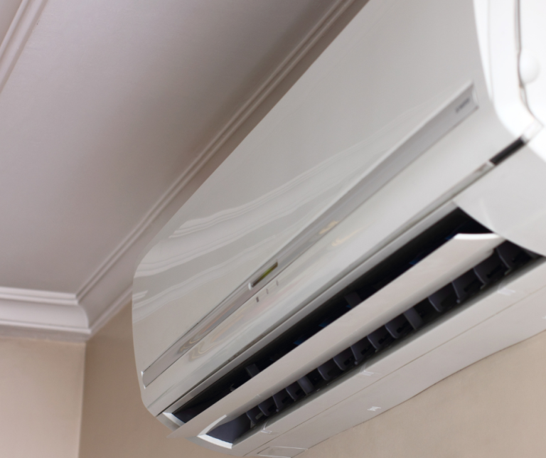 Close-up view of a white indoor heat pump air handler mounted near a ceiling, showing adjustable airflow vents and clean modern design against cream-colored walls with crown molding.