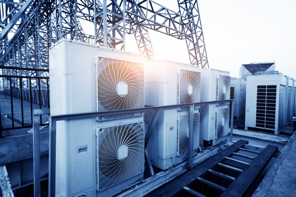 A row of industrial HVAC units on a rooftop with metal structures and the sun shining in the background.