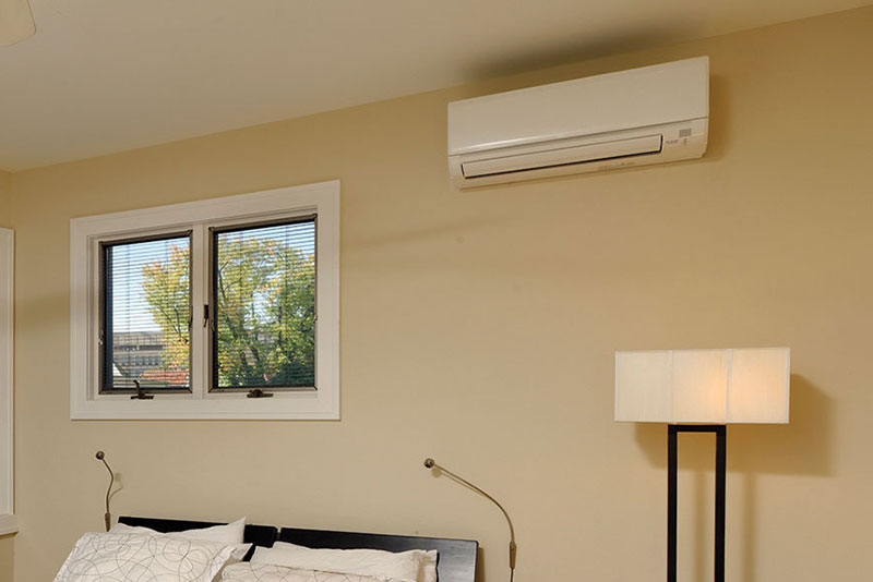 Wall-mounted mini-split air conditioner in a bedroom with a bed, window, and lamp.