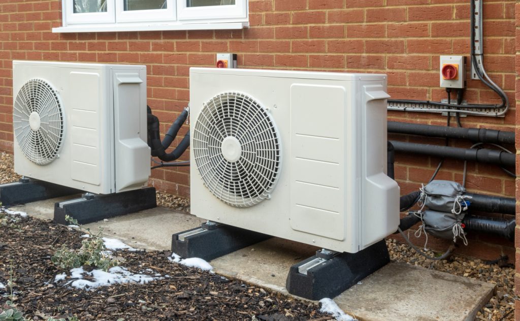Two outdoor heat pump units on raised platforms outside a brick wall, connected with pipes and wiring for heating and cooling.